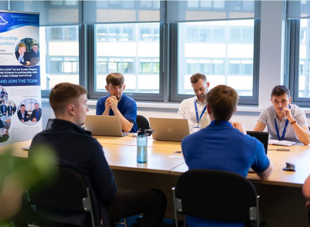 Five people sit around a table with laptops and water bottles, engaged in discussion in a bright office meeting room. A promotional banner for an engineering consultancy is visible in the background.