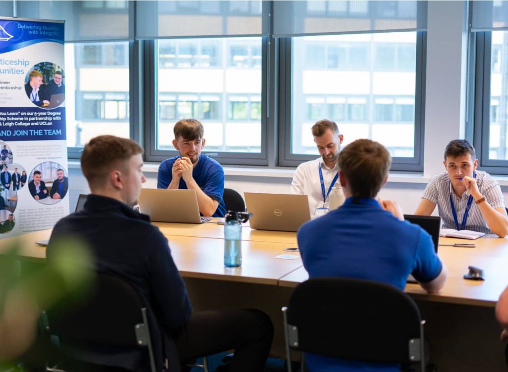 Five men wearing lanyards sit around a table with laptops, discussing engineering apprenticeships in a brightly lit office meeting room. A promotional banner stands in the background.