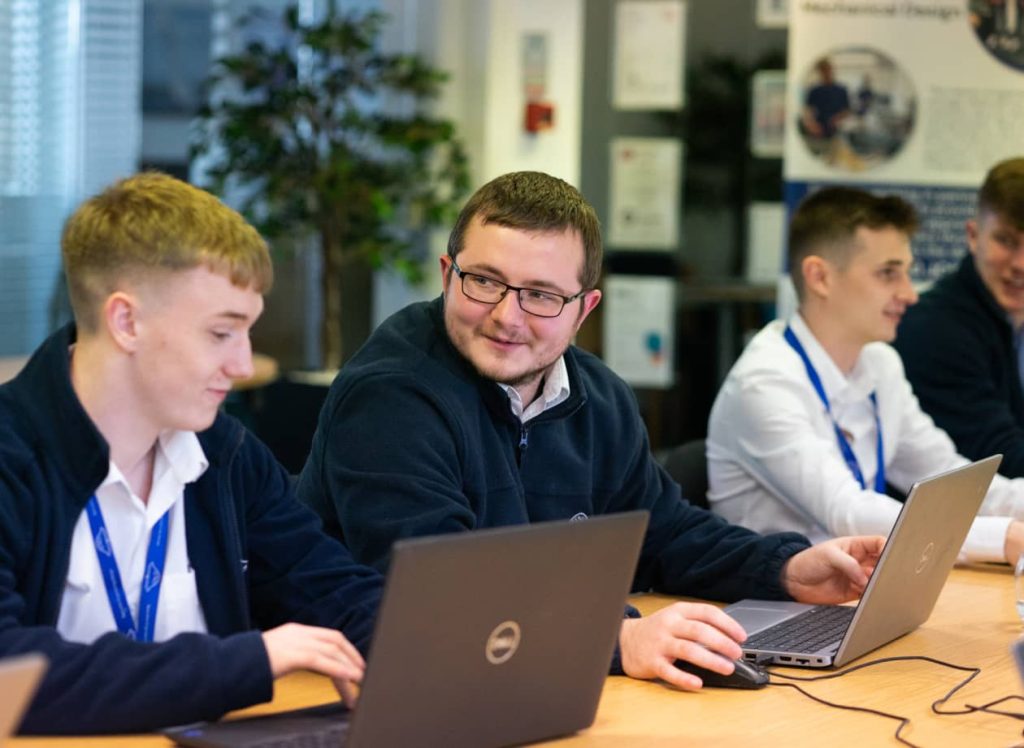 Four young men sit at a table working on laptops, wearing blue lanyards and casual business attire in an office setting, collaborating on engineering apprenticeships projects.