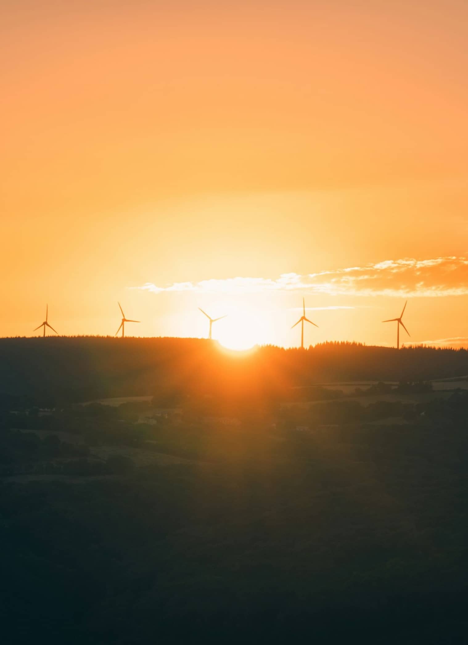 The sun sets behind a row of wind turbines on a ridgeline, casting an orange glow over the landscape—a striking symbol of nuclear sustainability and clean energy for the future.