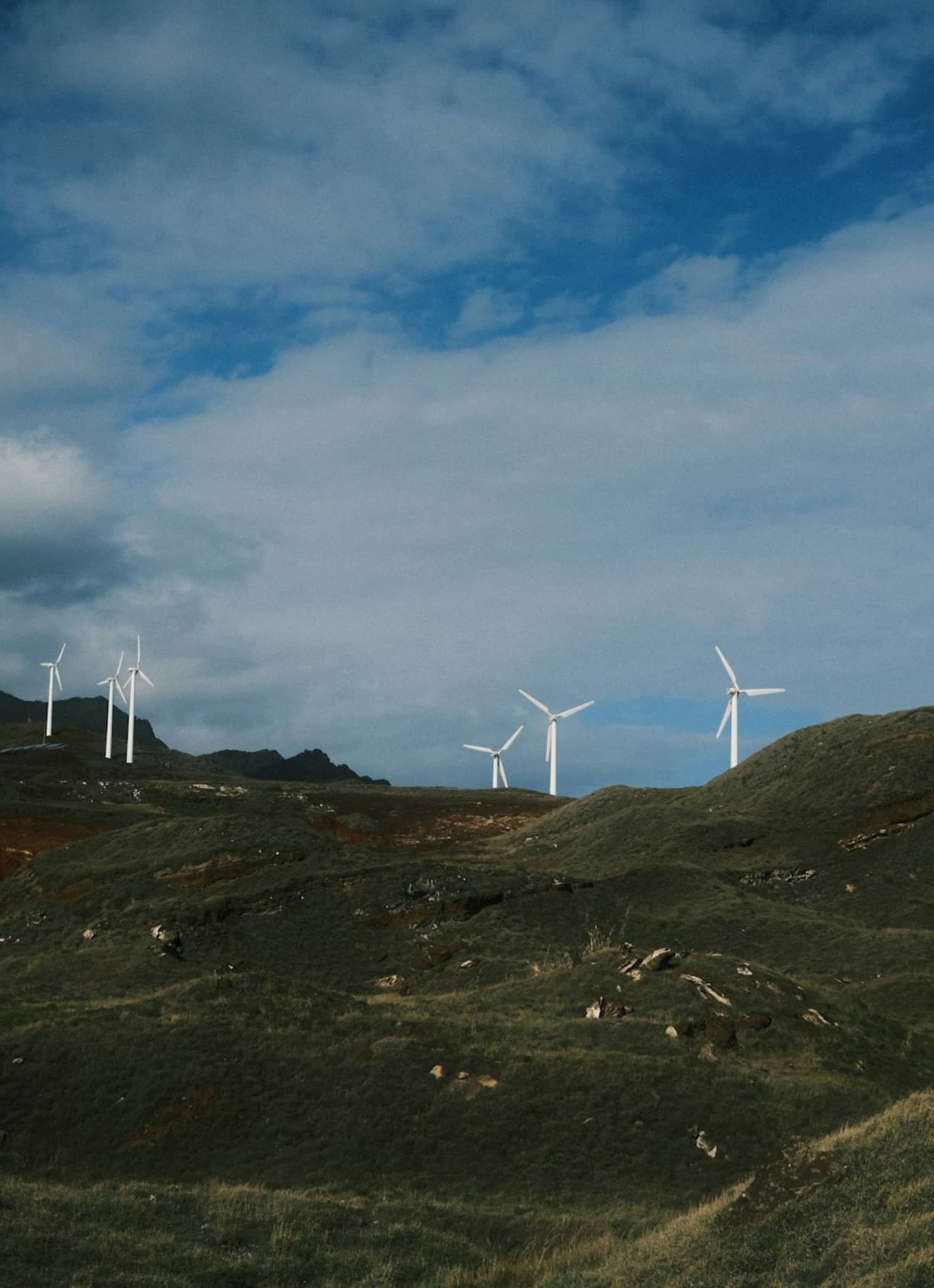 Wind turbines stand on grassy hills under a partly cloudy sky, generating renewables and green energy in a rural landscape.