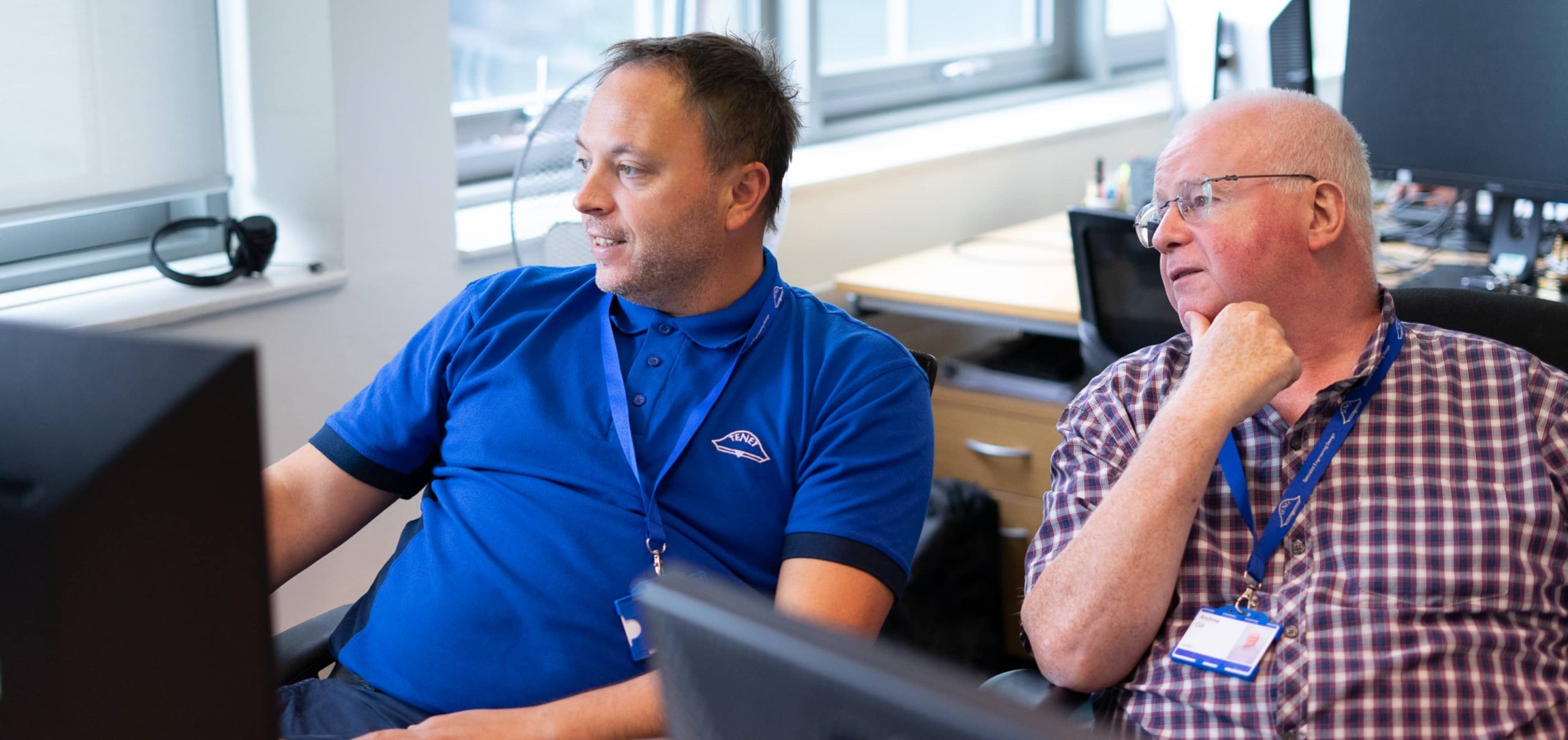 Two men from the Tenet Consultants team sit at desks in an office, looking at a computer screen and discussing something. Both wear lanyards with ID badges.